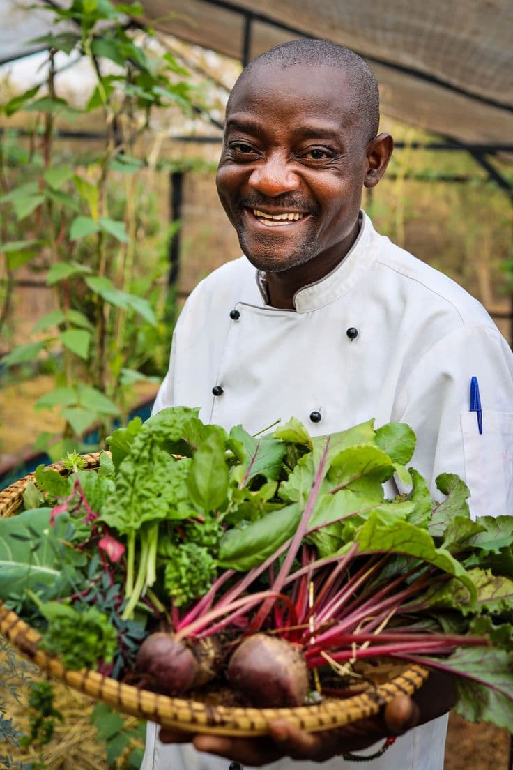 Smiling Black chef in a white coat holding a basket of fresh beets and greens.